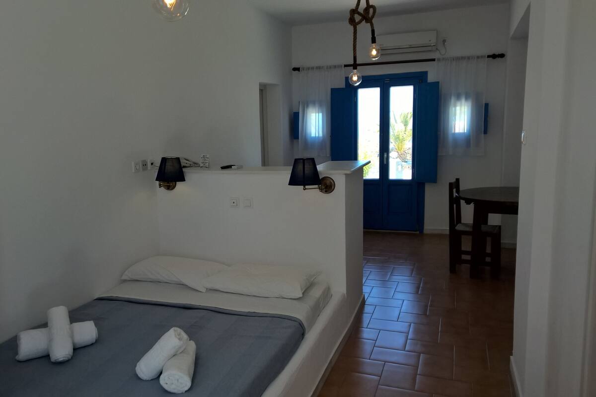 Une chambre d’hôtel avec un lit, un bureau et une chaise. Il y a une fenêtre avec des volets bleus et une porte qui donne sur l’extérieur.