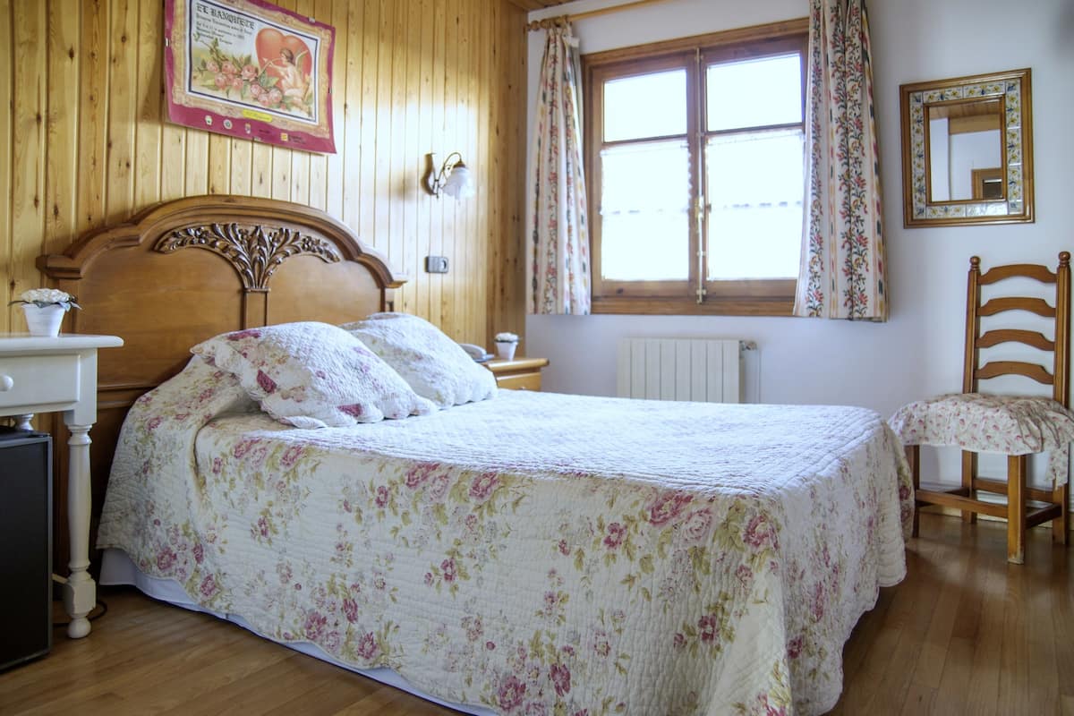 Un dormitorio con cabecera de madera, colcha estampada con motivos florales, una mesita de noche, una silla y una ventana con cortinas.