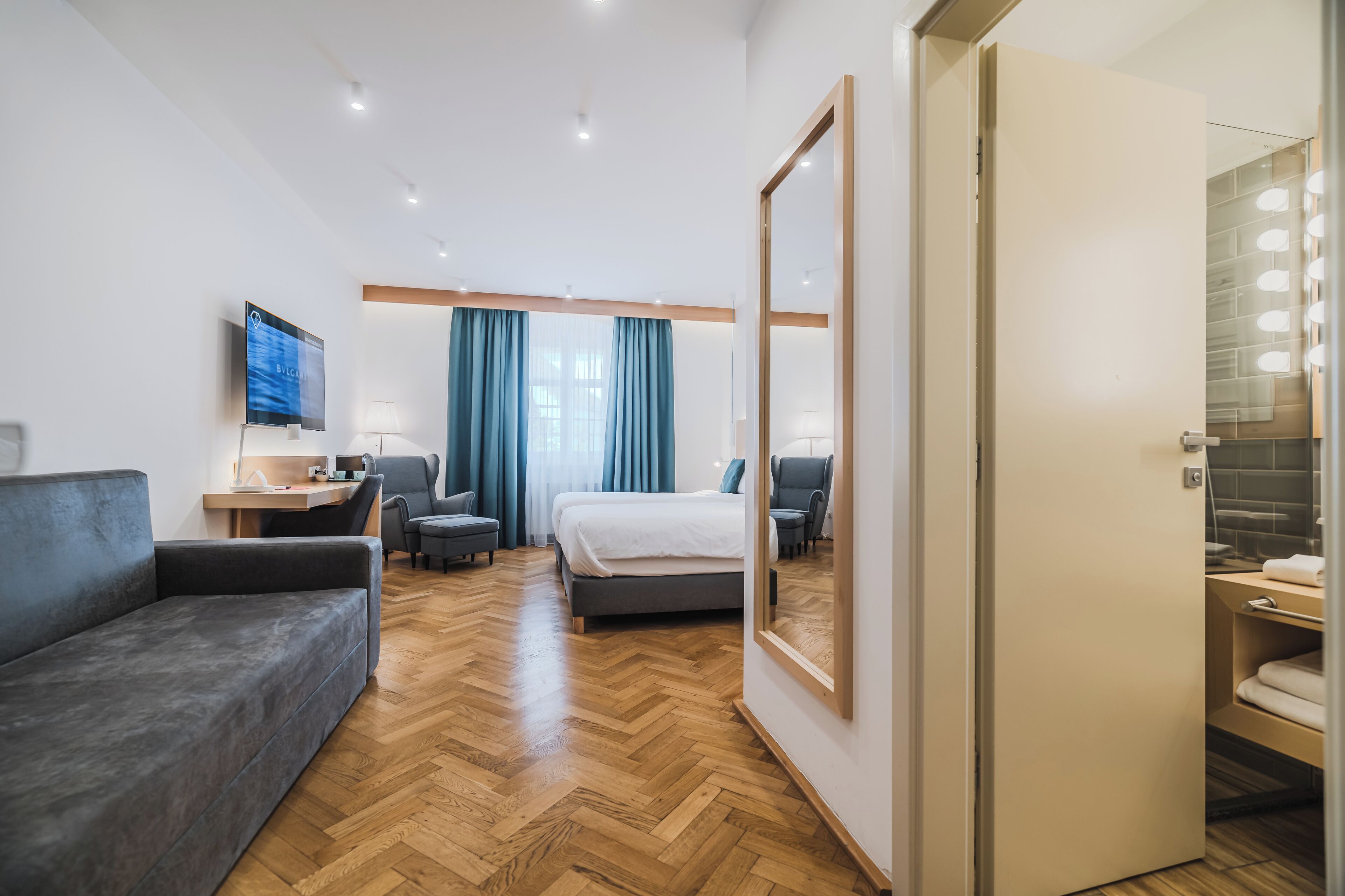 Ein modernes Hotelzimmer mit Bett, Sofa, Schreibtisch, Fernseher und einem Badezimmer mit Spiegel und Beleuchtung.