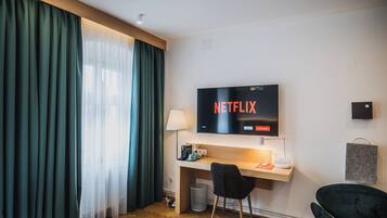 Hotelkamer met een bed, bureau, stoel, tv waarop 'NETFLIX' wordt weergegeven, en een raam met gordijnen.