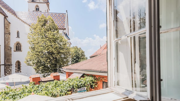 A room with a large window overlooking a courtyard, a wooden table, and a view of a church with a spire. - Casa Luxemburg (Sibiu)
