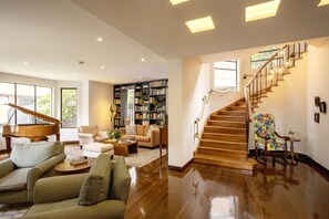 A spacious living room with a grand piano, a bookshelf, and a staircase with a decorative chair. - Bh la Quinta- Suites (Bogotá)