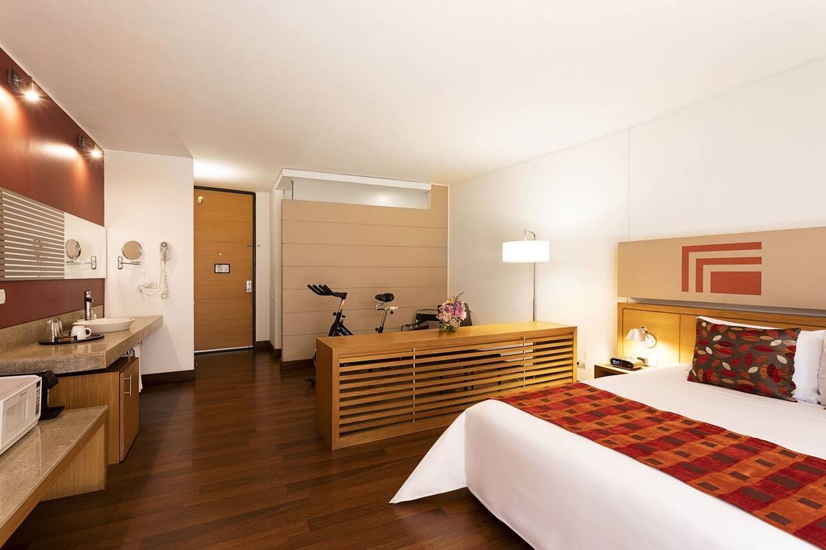 Ein Hotelzimmer mit einem Holztisch, einem Bett mit gemustertem Überwurf, einer Nachttischlampe und einem großen Spiegel.