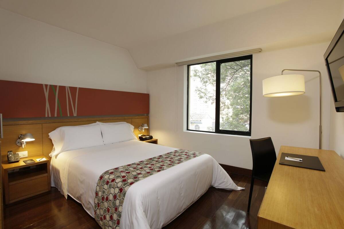 Ein Hotelzimmer mit Bett, Nachttischen, Schreibtisch, Stuhl, Lampe und Fenster mit Blick.
