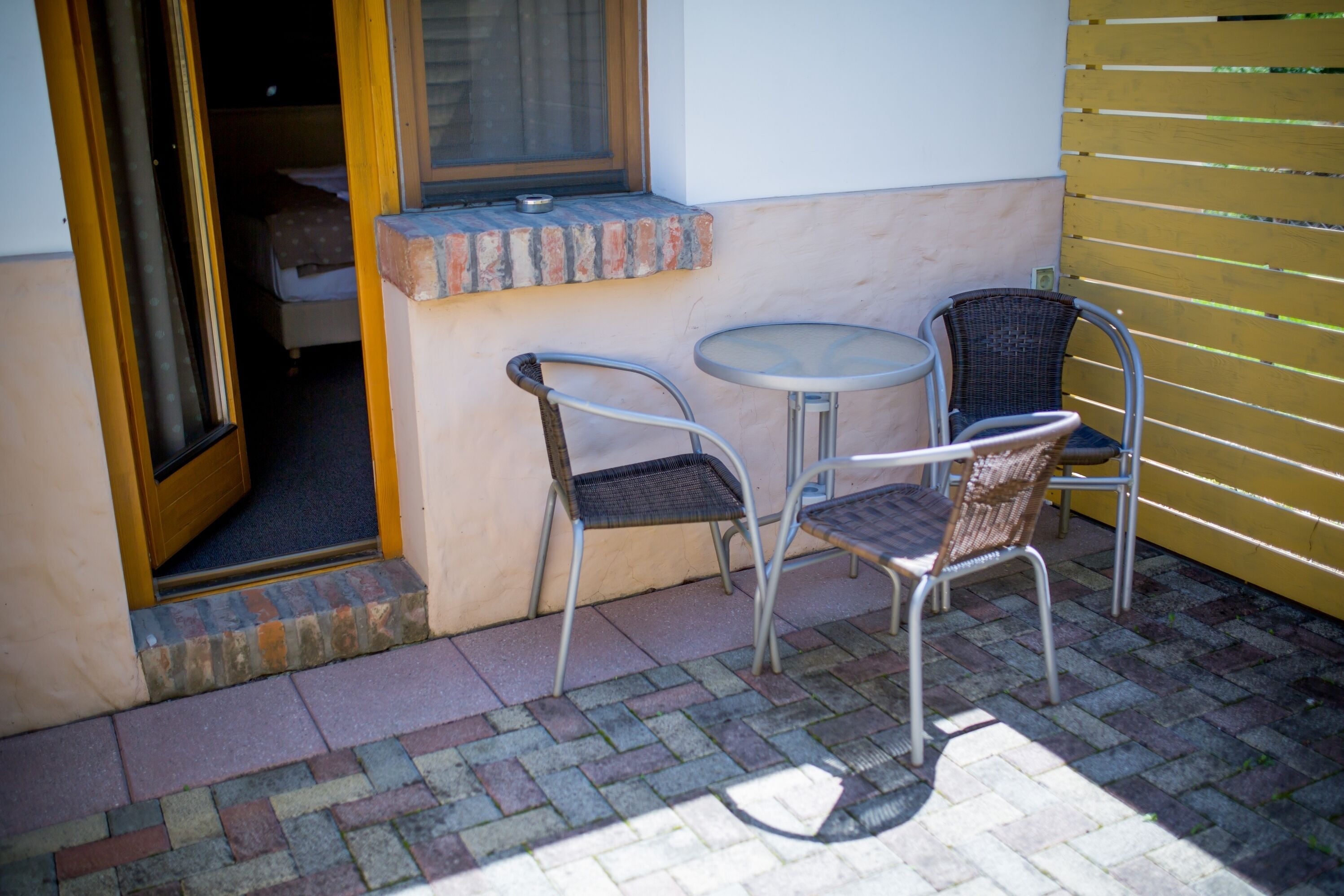 Ein Sitzbereich im Freien mit zwei Metallstühlen und einem kleinen runden Tisch, neben einem Gebäude mit Fenster und Backsteinschräge.