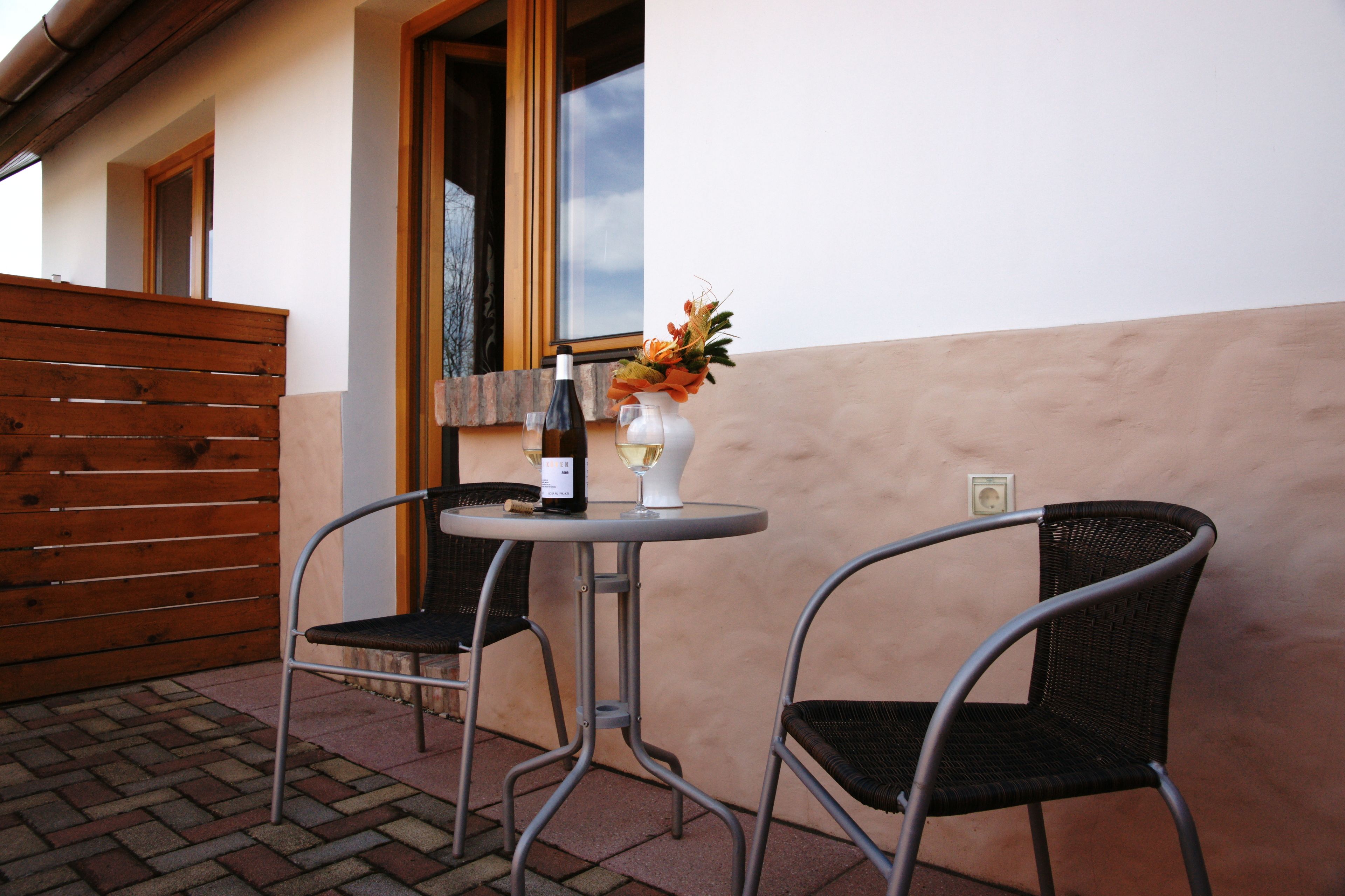 Een terras met twee metalen stoelen, een tafel, een fles wijn en een vaas met bloemen.