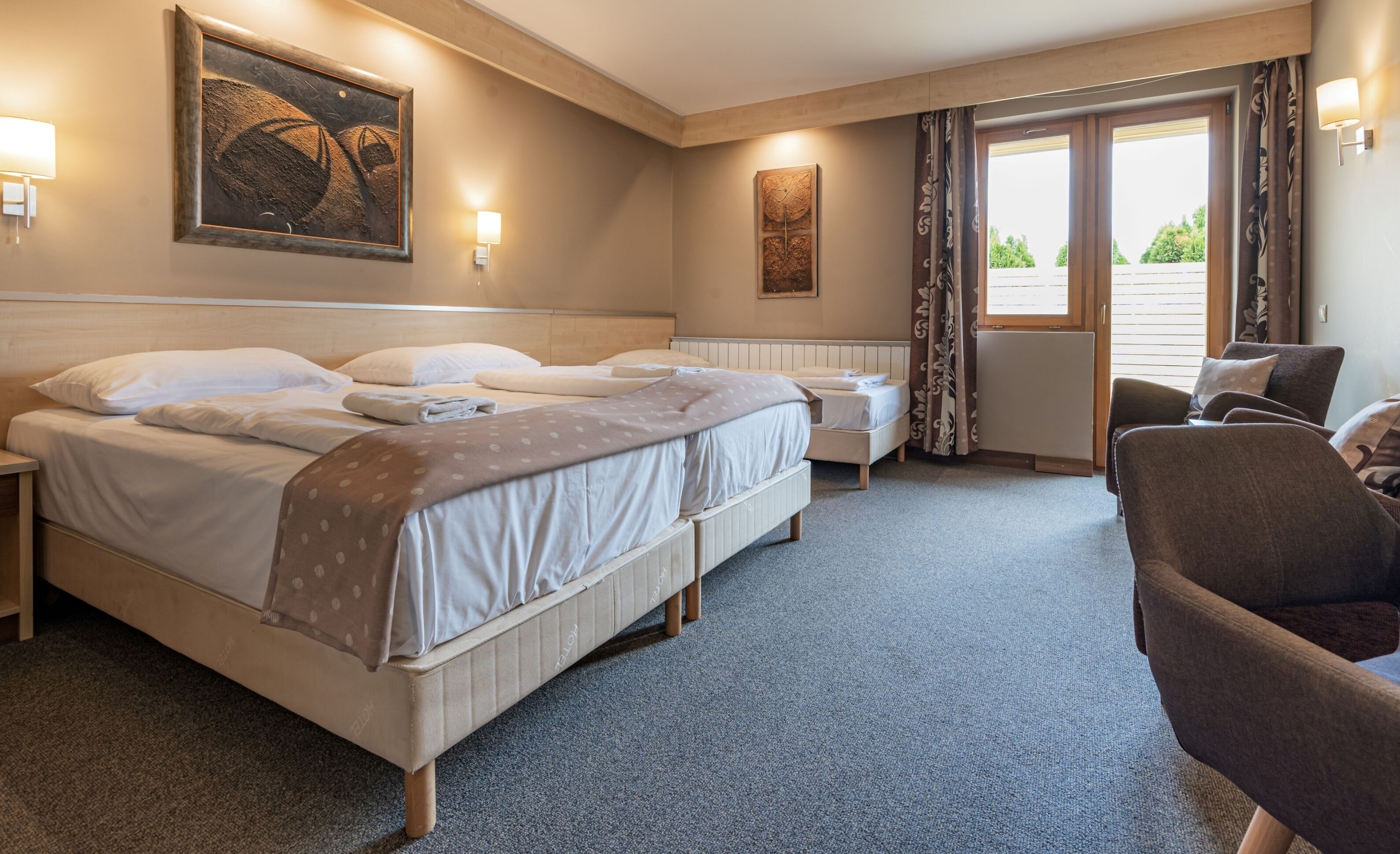 A hotel room with a large bed, a smaller bed, a sofa, and a desk. There are two wall-mounted lamps and a window with curtains.