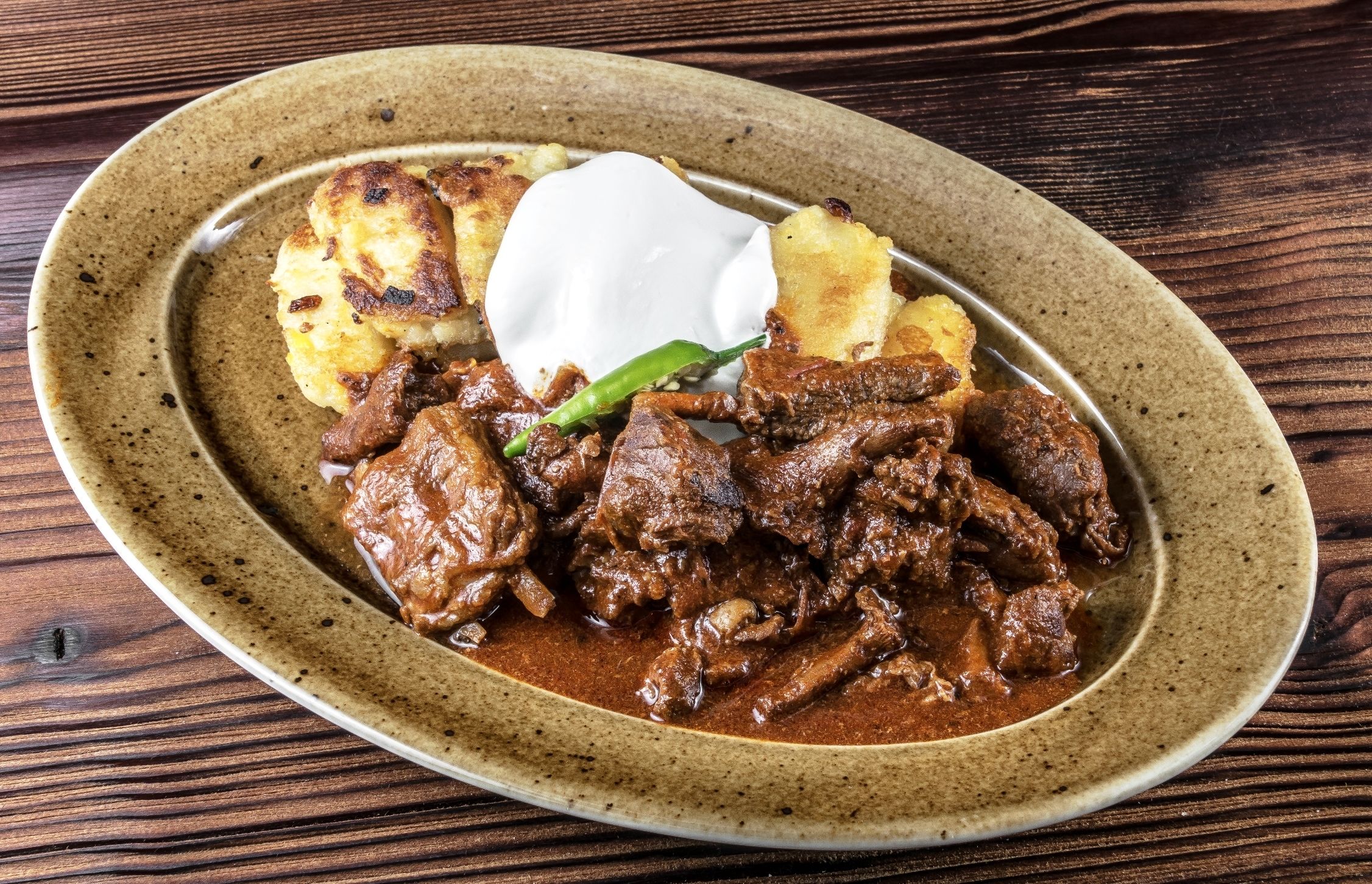A plate of food with meat and potatoes, a dollop of sour cream, and a green chili.