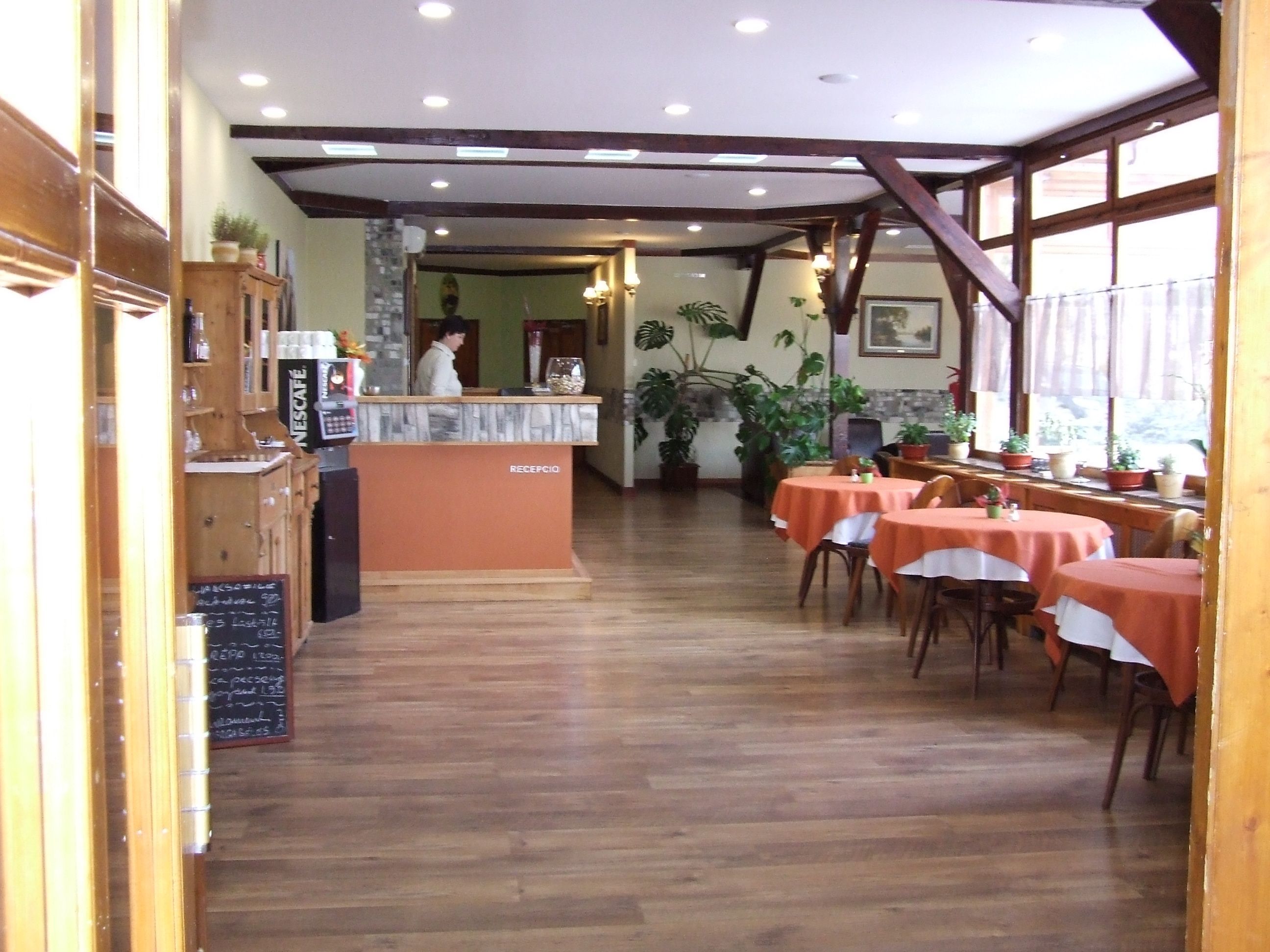 Een restaurant interieur met een receptiebalie, houten vloer, tafels met oranje tafelkleden en potplanten.