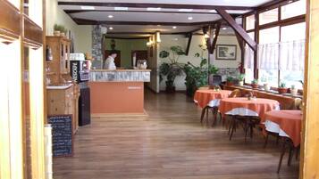 Een restaurant interieur met een receptiebalie, houten vloer, tafels met oranje tafelkleden en potplanten.