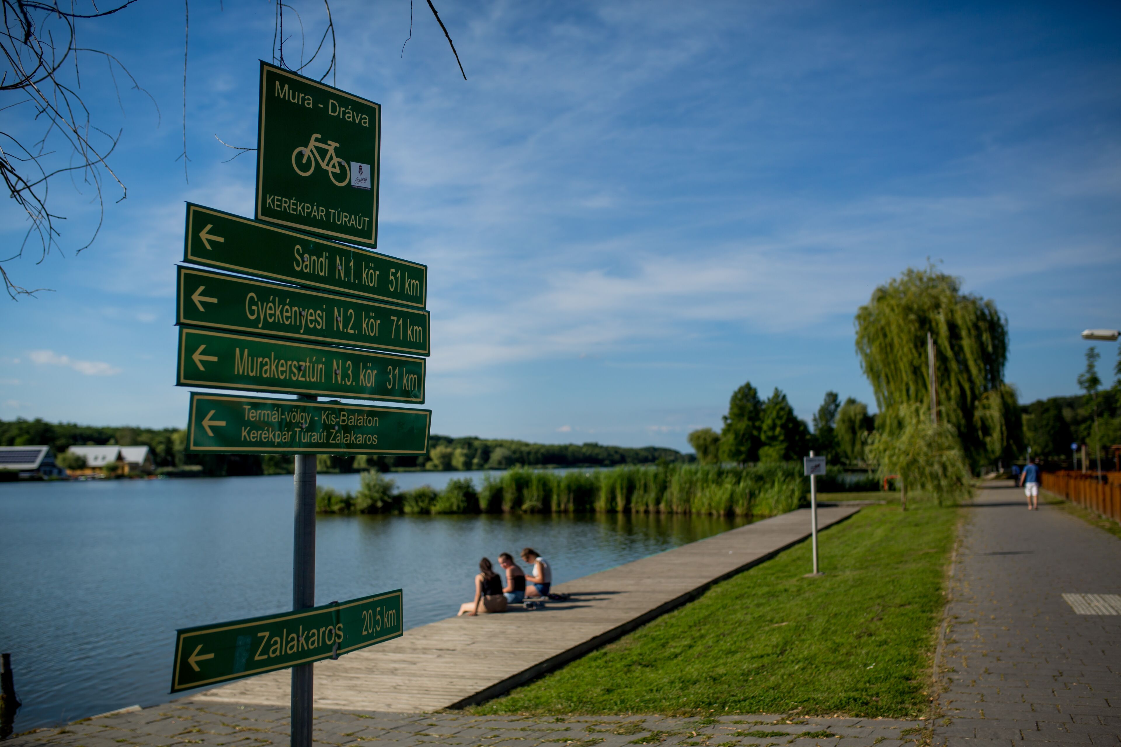 Ein Wegweiser am Ufer eines Flusses, mit Menschen, die auf dem Pfad sitzen.