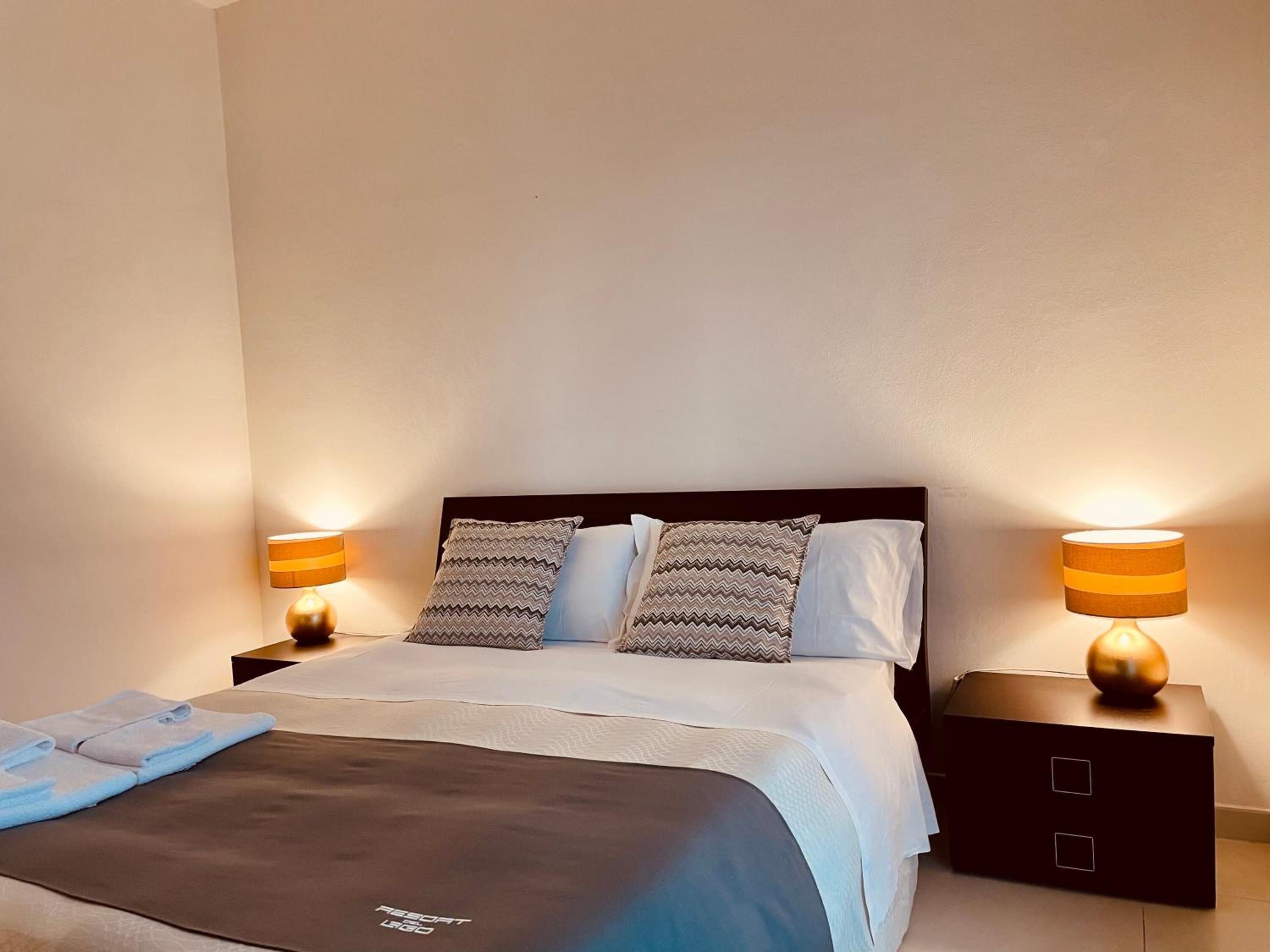 A neatly made bed with a dark headboard, two patterned pillows, and a matching blanket. There are two bedside tables with lamps, and a small box on the right side of the bed.