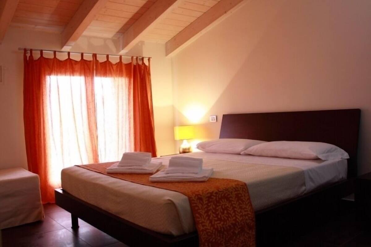 Una camera d'albergo con un letto, un comodino, una lampada e una finestra con tende.