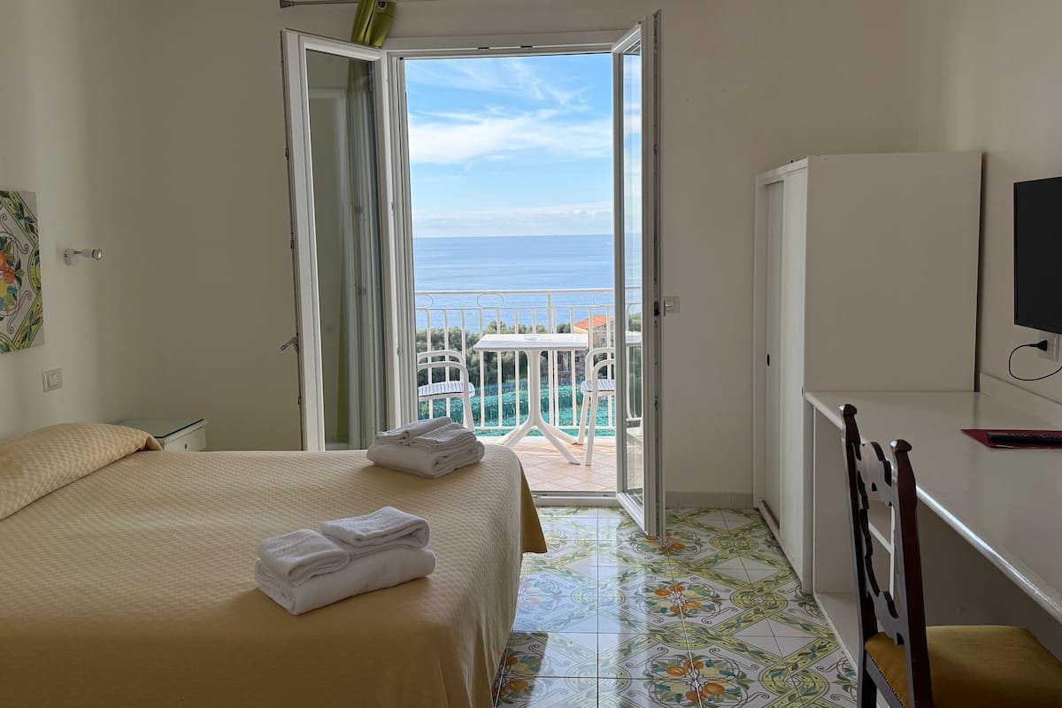 Ein Hotelzimmer mit Bett, Schreibtisch, Fernseher und einem Balkon mit Meerblick.