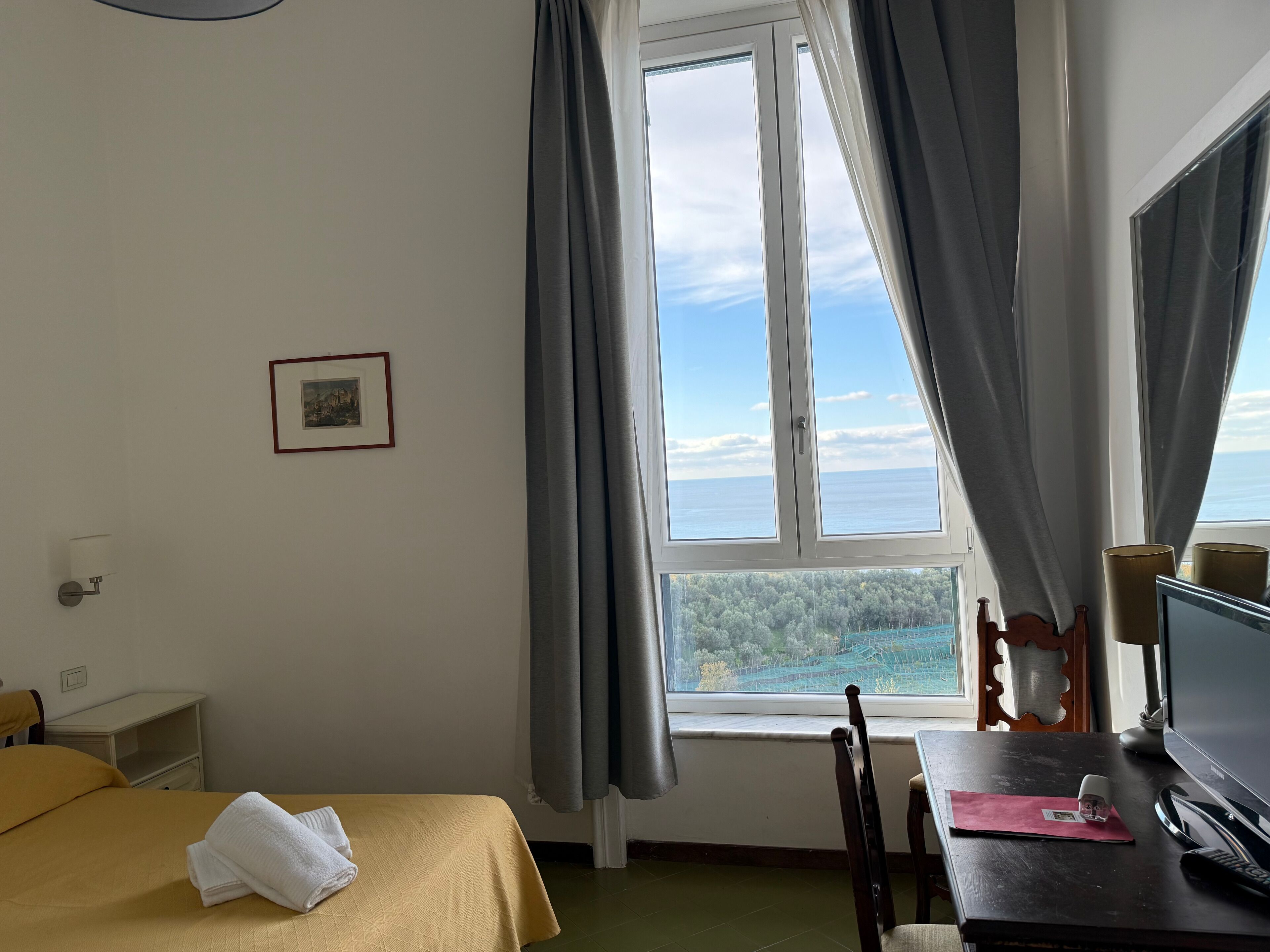 A hotel room with a bed, a desk, a chair, a window with curtains, a framed picture on the wall, and a view of the sea and trees.