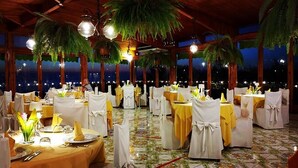 An indoor dining area with round tables covered in yellow tablecloths and white chairs, set against a backdrop of large windows and palm trees. - Hotel Villa Pina Antico Francischiello (Massa Lubrense)