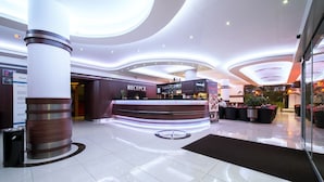 A hotel reception area with a modern design, featuring a reception desk, a large pillar, and a seating area with potted plants. - Alessandria (Hradec Kralove)