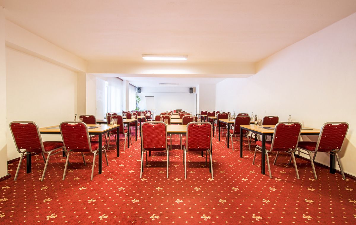 A conference room with multiple tables and chairs, carpeted floor, and a plain white wall.