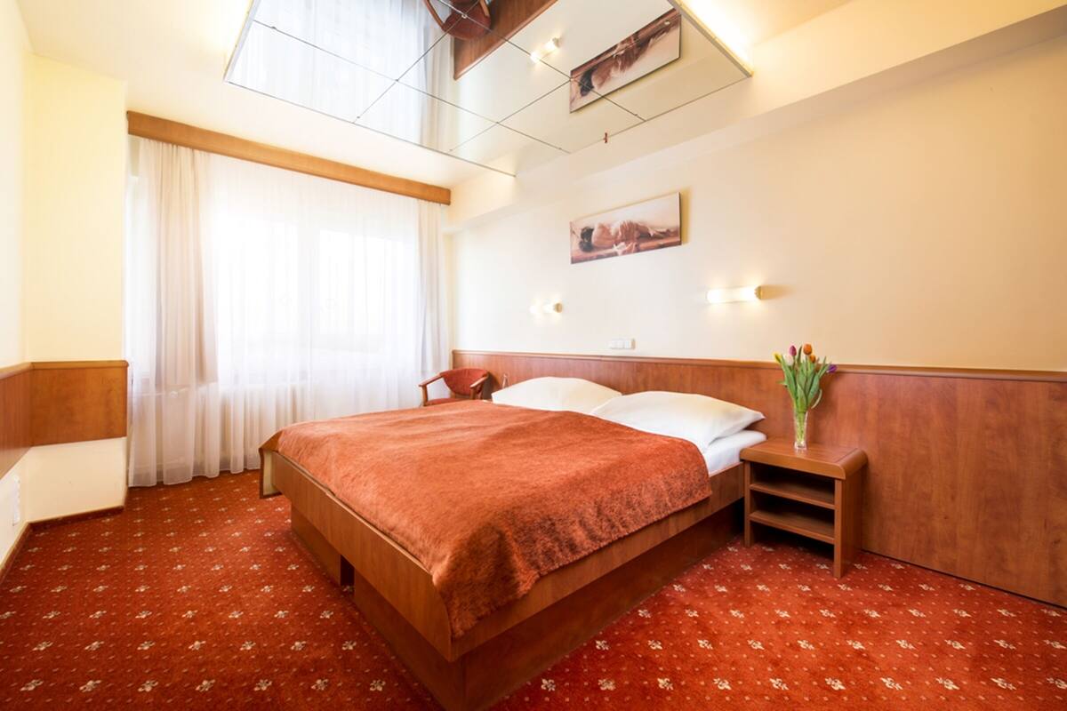 A hotel room with a large bed, a wooden headboard, a bedside table with a vase of flowers, and a window with sheer curtains.