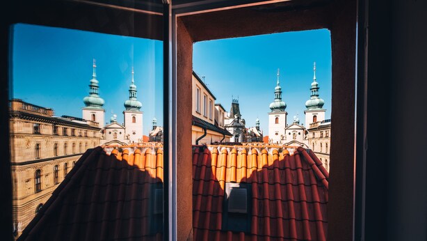 A view through a window with a red-tiled roof, looking out at a building with multiple domed towers and a clear blue sky. - Pension Corto (Prague)