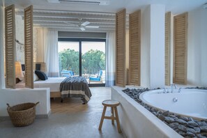 A bedroom with a large window, a bed, a wooden stool, a bathtub, and a balcony with chairs.