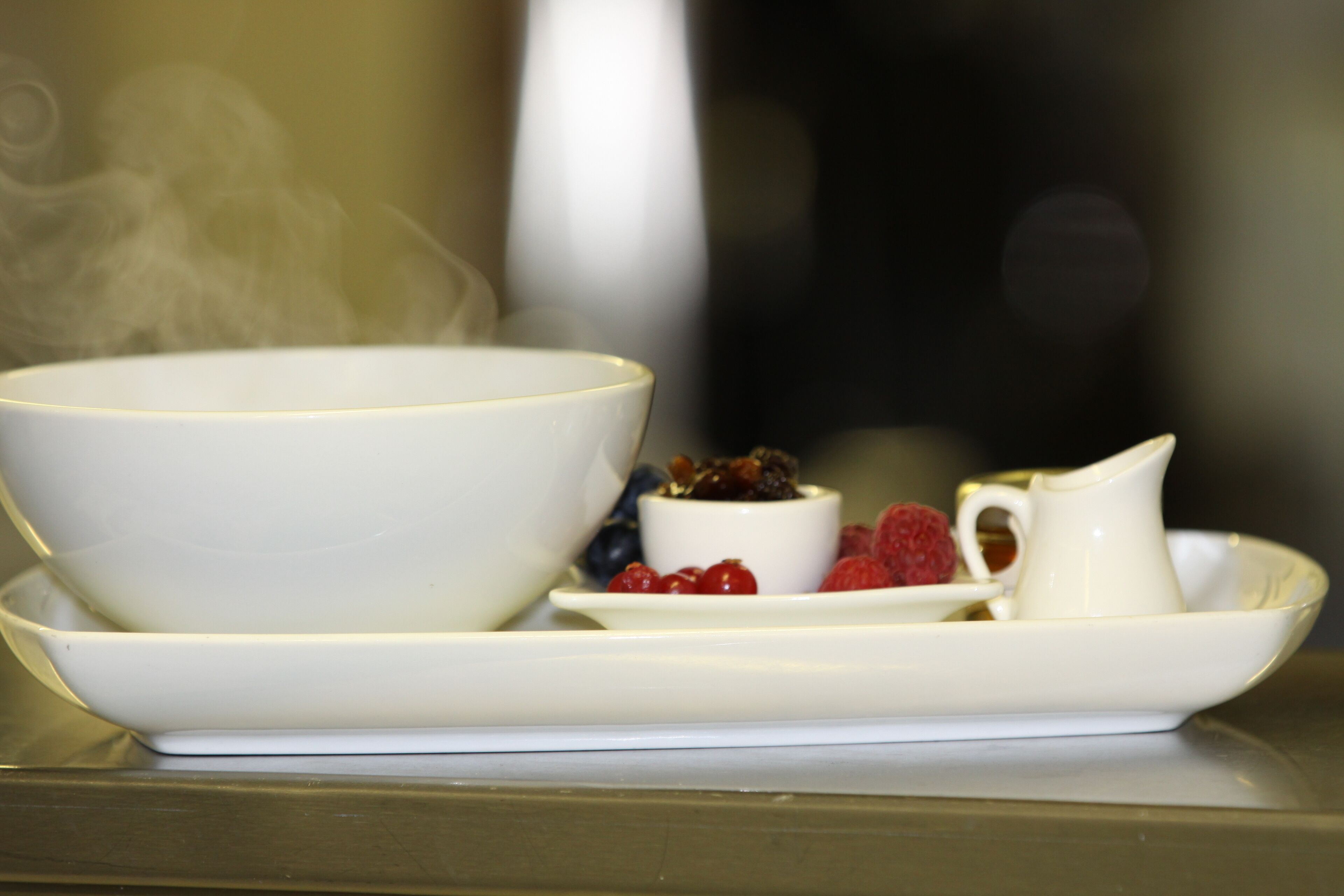 A white bowl, a small cup, and a pitcher on a white plate with berries and a slice of fruit.