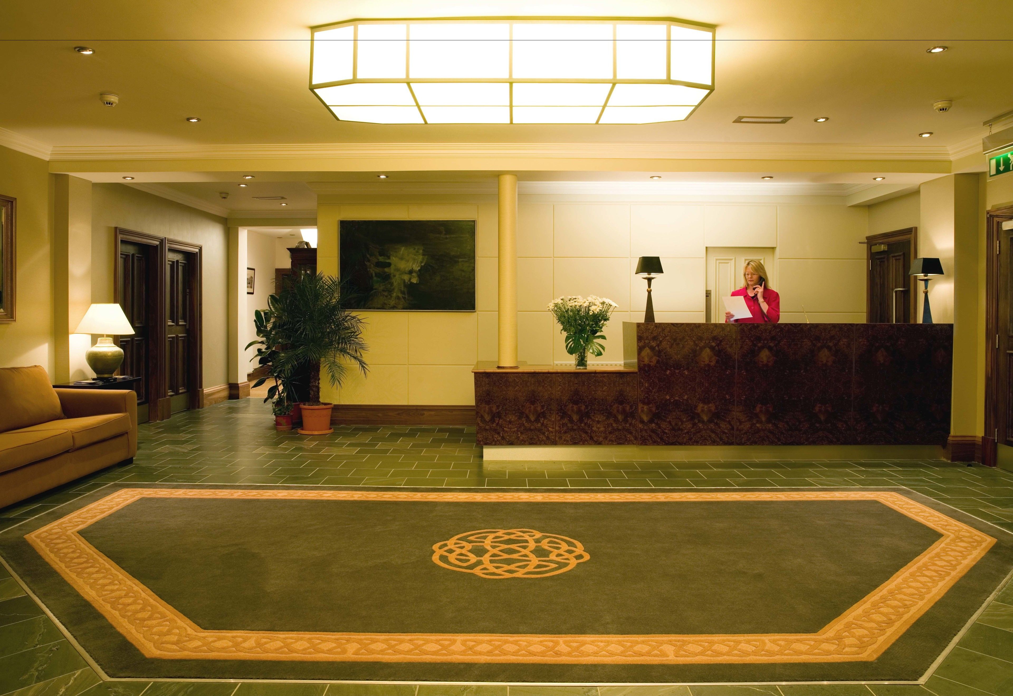 A hotel reception area with a reception desk, a large decorative rug, a sofa, and a potted plant.