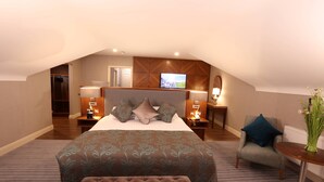 Quarto de hotel com uma cama grande, um espaço de estar com uma cadeira e uma mesa, uma televisão e uma pequena escrivaninha com uma luminária.