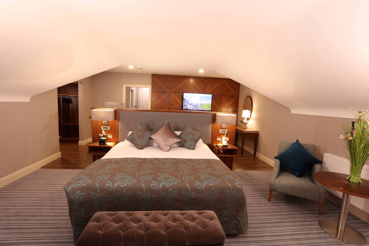 A hotel room with a large bed, a sitting area with a chair and a table, a television, and a small desk with a lamp.
