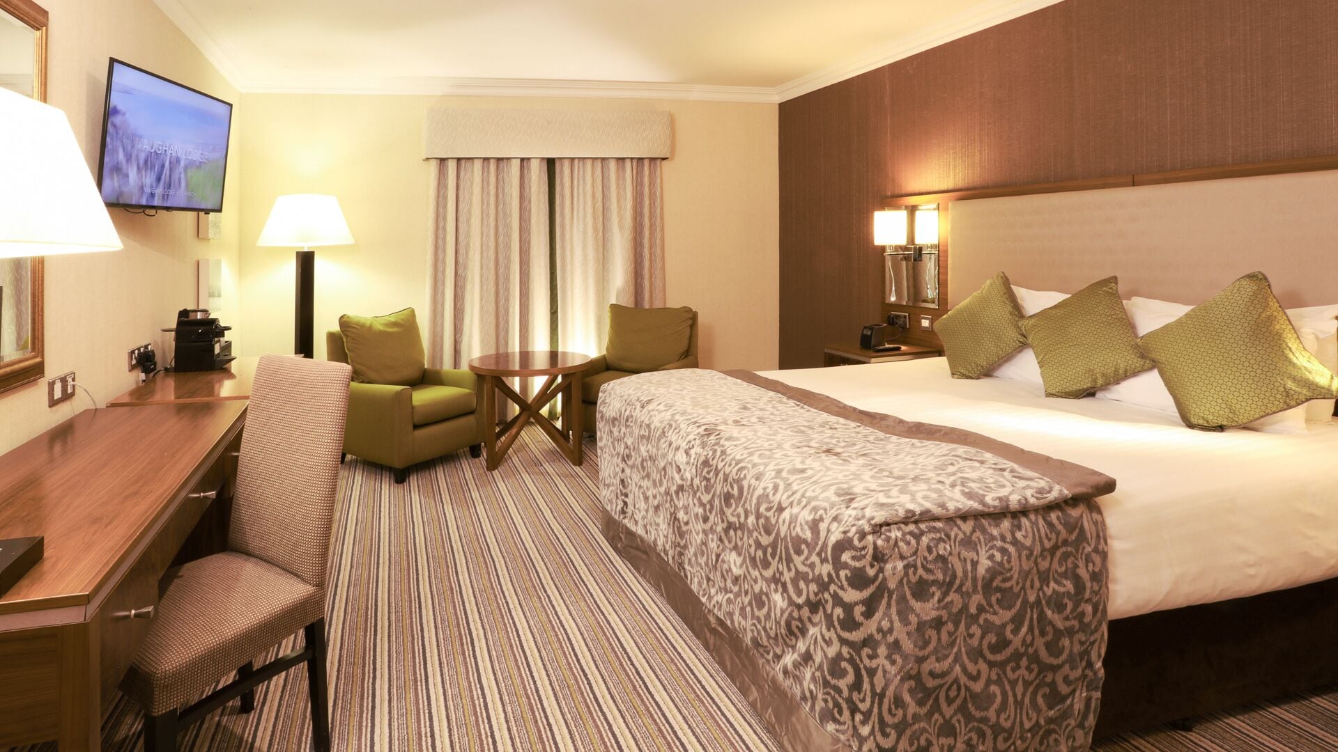 A hotel room with a bed, a desk, a chair, a small table, a TV, and a window with curtains.
