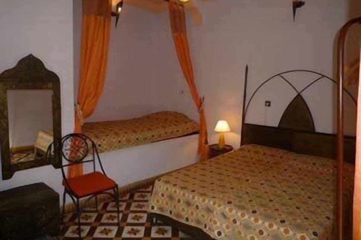 Un dormitorio con una cama individual, una silla, una mesita de noche con lámpara y un espejo en la pared.