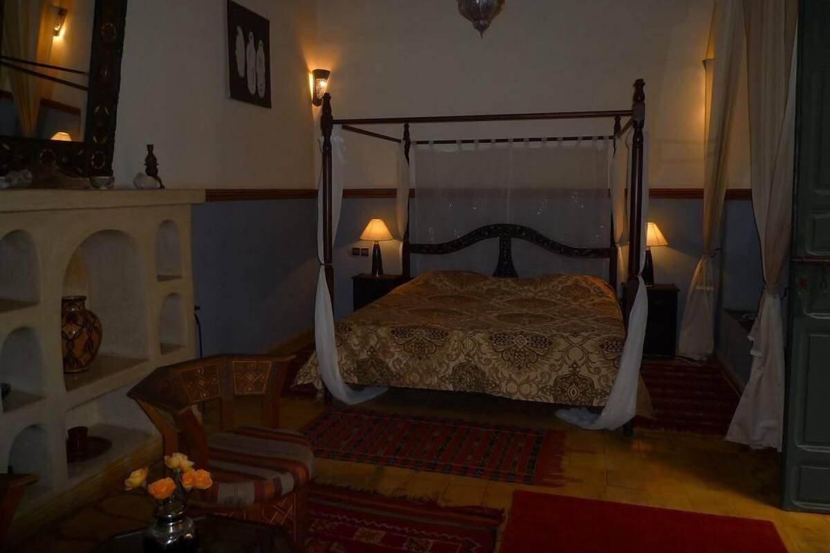 Una cama con dosel, con una colcha estampada, dos lámparas de noche, un florero con flores y una alfombra roja en el suelo.