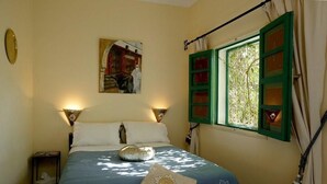 A bedroom with a bed, a nightstand, a window with green shutters, and a painting on the wall.