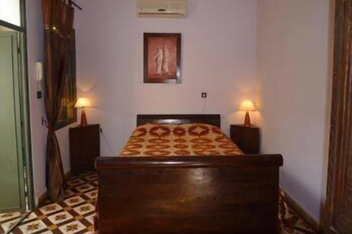 Un dormitorio con una cama, dos lámparas de noche, aire acondicionado de pared y una puerta con cortinas.