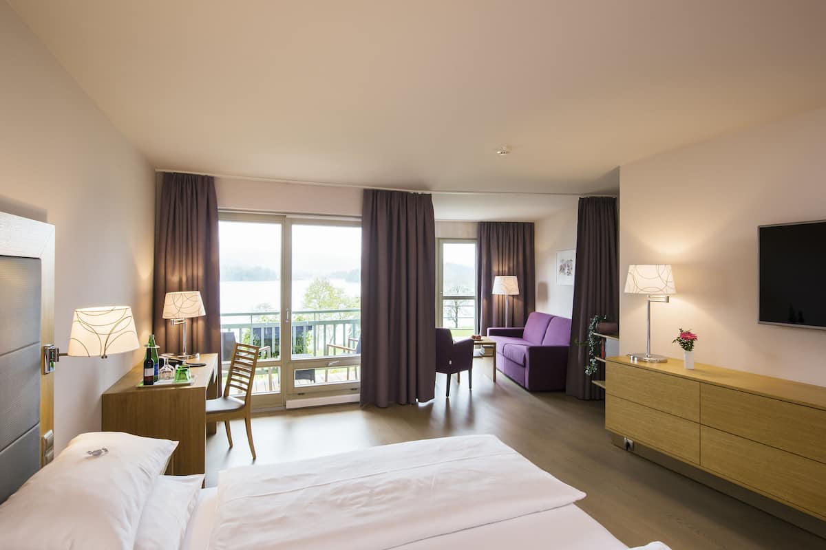 Una habitación de hotel con cama, escritorio con lámpara, silla, sofá, televisor y balcón con cortinas.