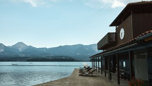 Un muelle de madera con sillones de descanso que se extiende sobre un lago, un edificio con un reloj y un veleta, y montañas al fondo.