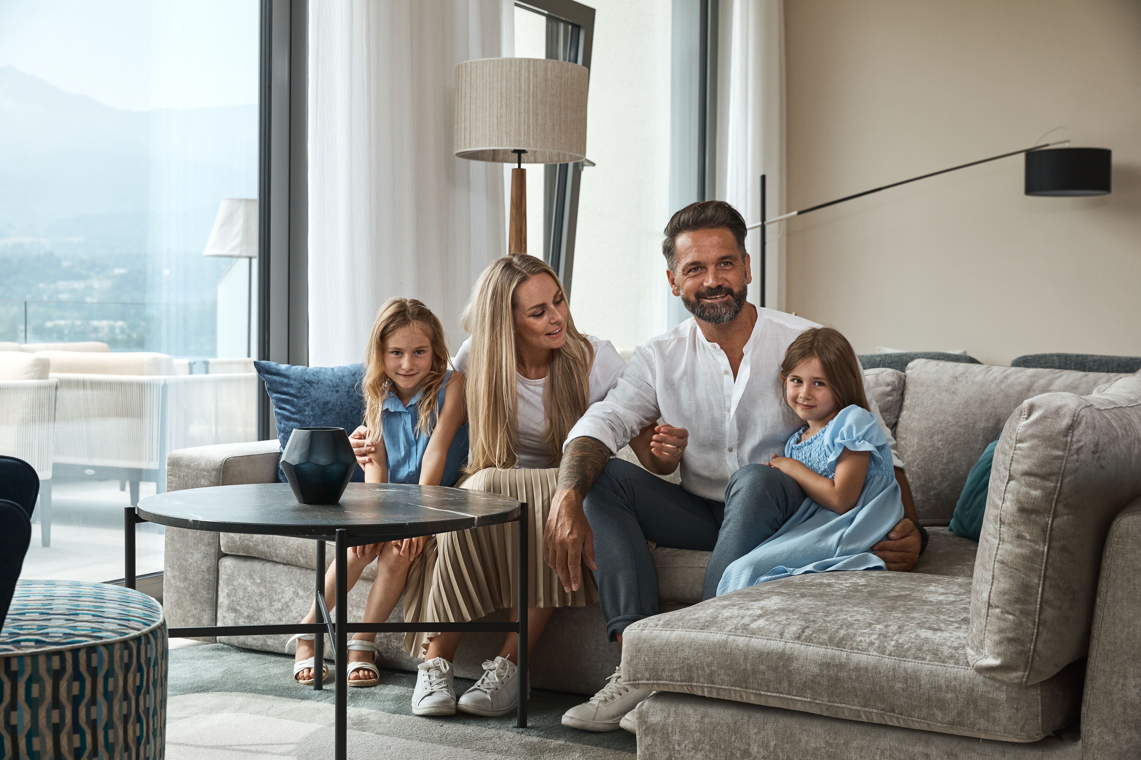 Eine Familie sitzt auf einem Sofa in einem gut beleuchteten Raum mit einem großen Fenster, einer Lampe und einem kleinen Tisch.