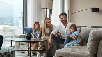 Eine Familie sitzt auf einem Sofa in einem gut beleuchteten Raum mit einem großen Fenster, einer Lampe und einem kleinen Tisch.