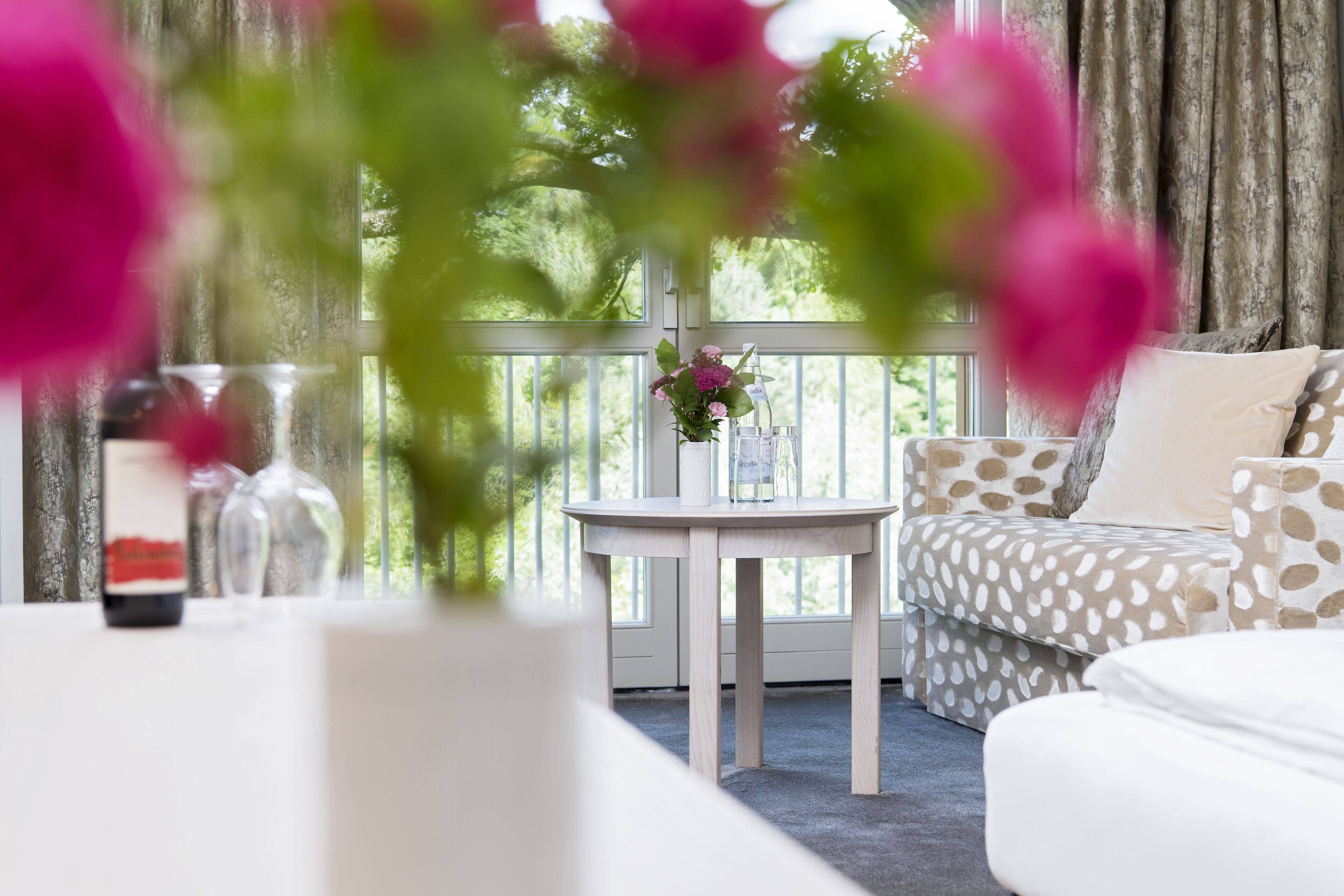 Ein Zimmer mit einem weißen Sofa, einem runden Tisch mit Blumen in einer Vase und Blick auf Grün durch ein Fenster.