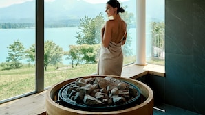 Una persona de pie junto a una ventana con vista a un lago y montañas al fondo.