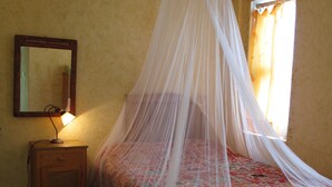 A bedroom with a bed under a mosquito net, a bedside table with a lamp, a mirror on the wall, and a patterned rug. - Daphne House (Kumluca)
