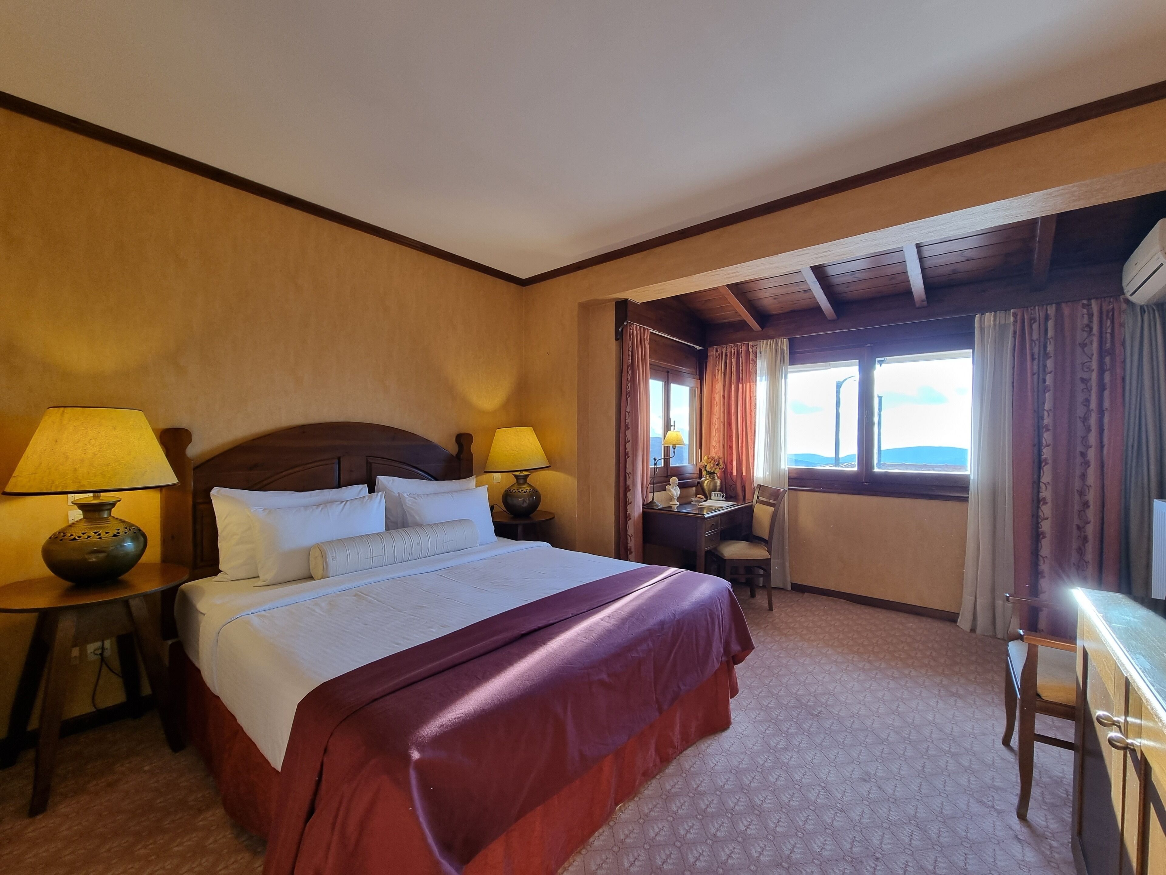 A hotel room with a large bed, two bedside lamps, a desk with a chair, and a window with curtains.
