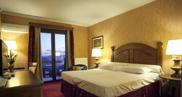 Habitación de hotel con cama, dos mesitas de noche con lámparas, un escritorio con silla, una ventana con cortinas y vistas a las montañas.