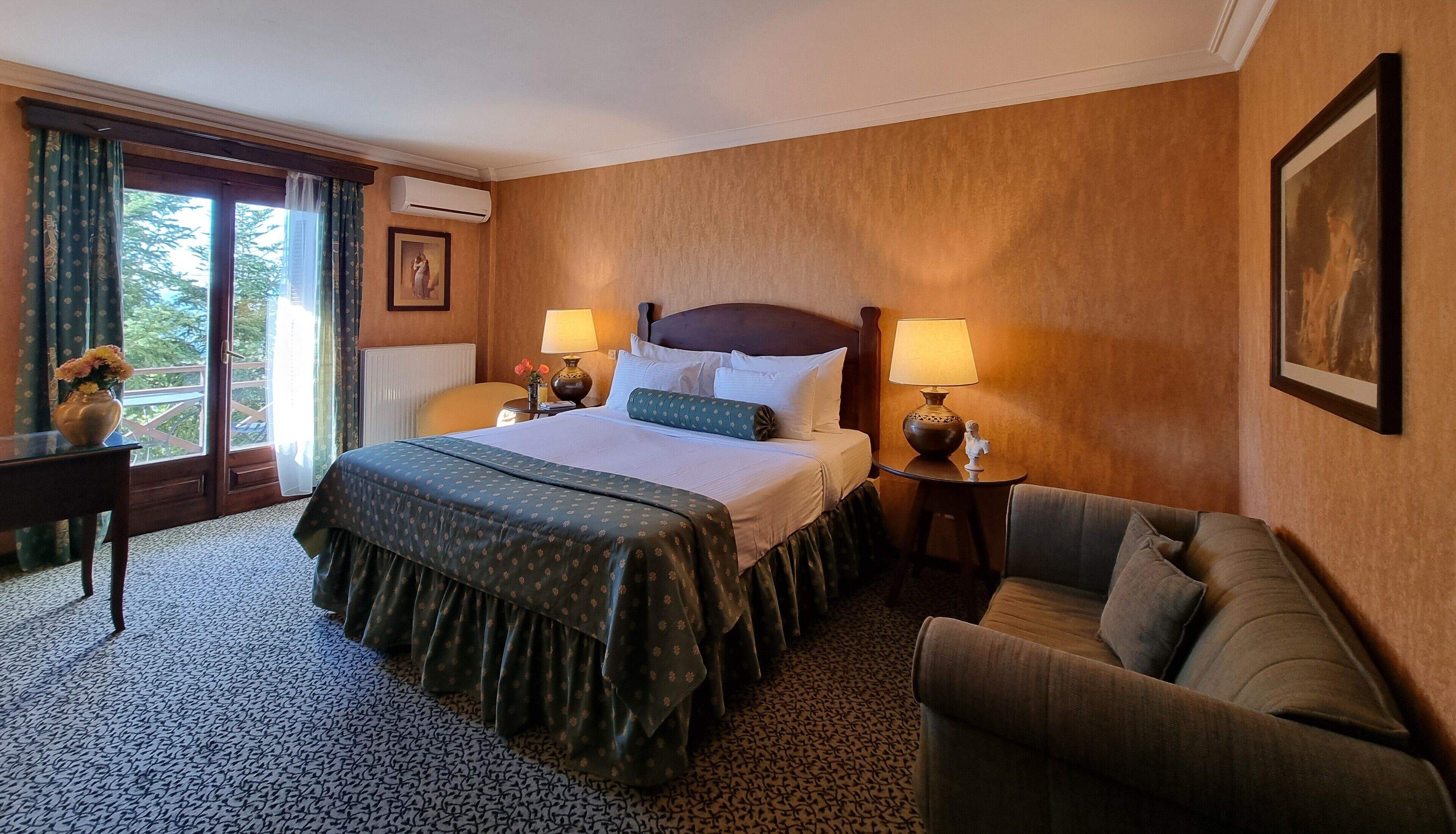 A hotel room with a large bed, two bedside tables with lamps, a sofa, a window with curtains, and a framed picture on the wall.