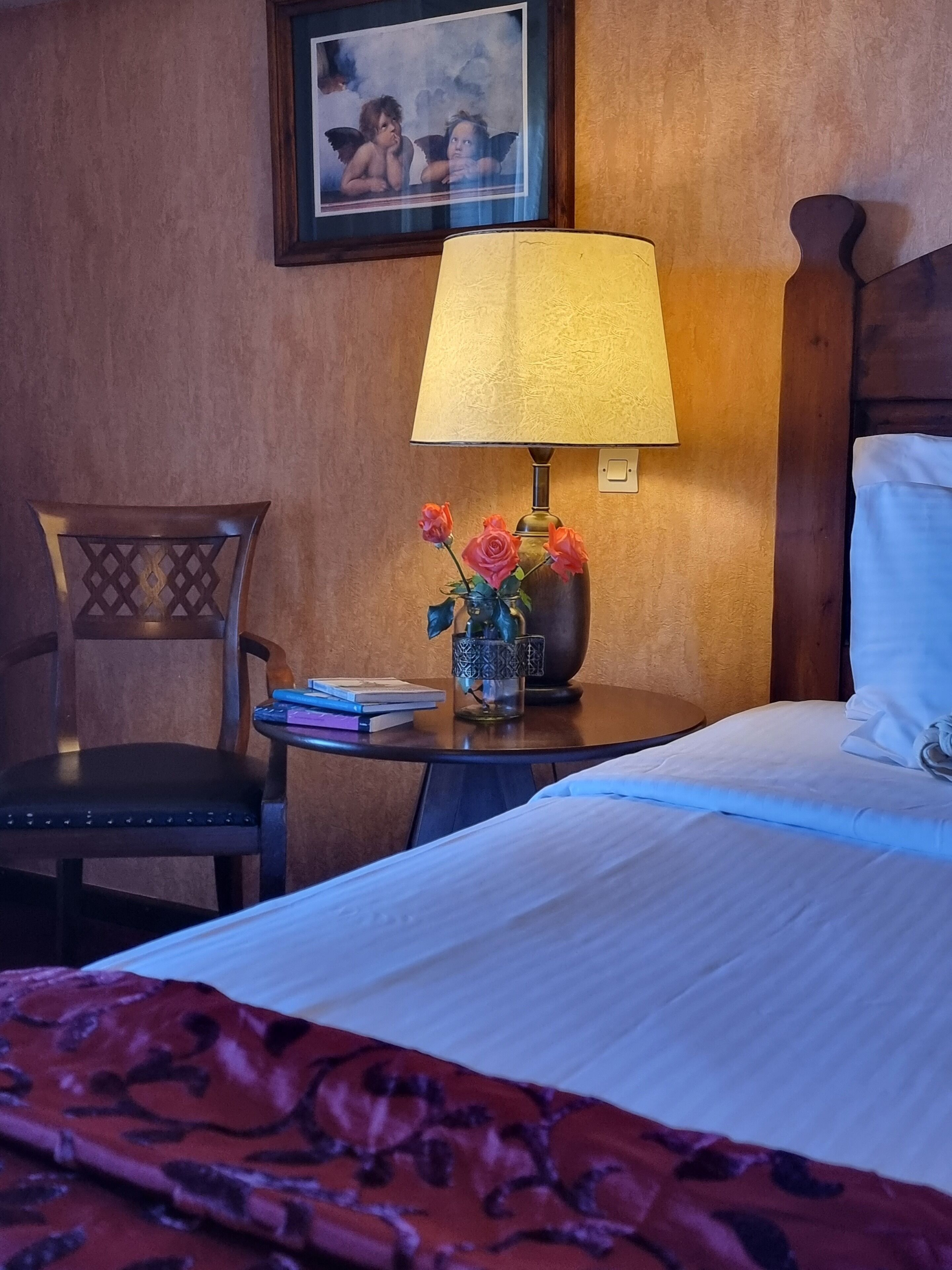 A hotel room with a bed, a nightstand with a lamp, a vase with flowers, a chair, and a framed picture on the wall.