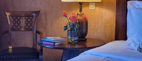 A hotel room with a bed, a nightstand with a lamp, a vase with flowers, a chair, and a framed picture on the wall.