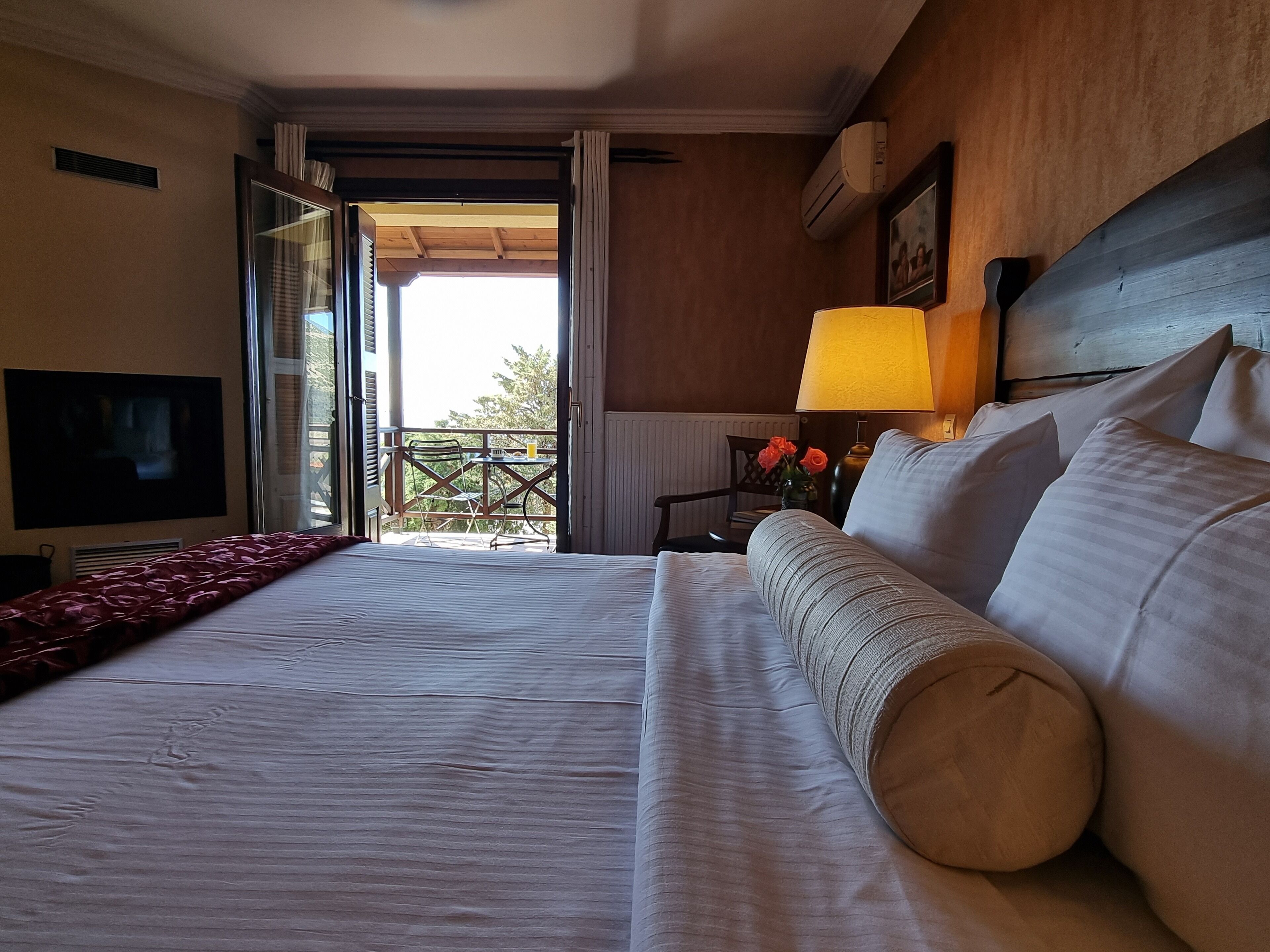 A hotel room with a large bed, a TV, a balcony with a table and chairs, and a lamp.