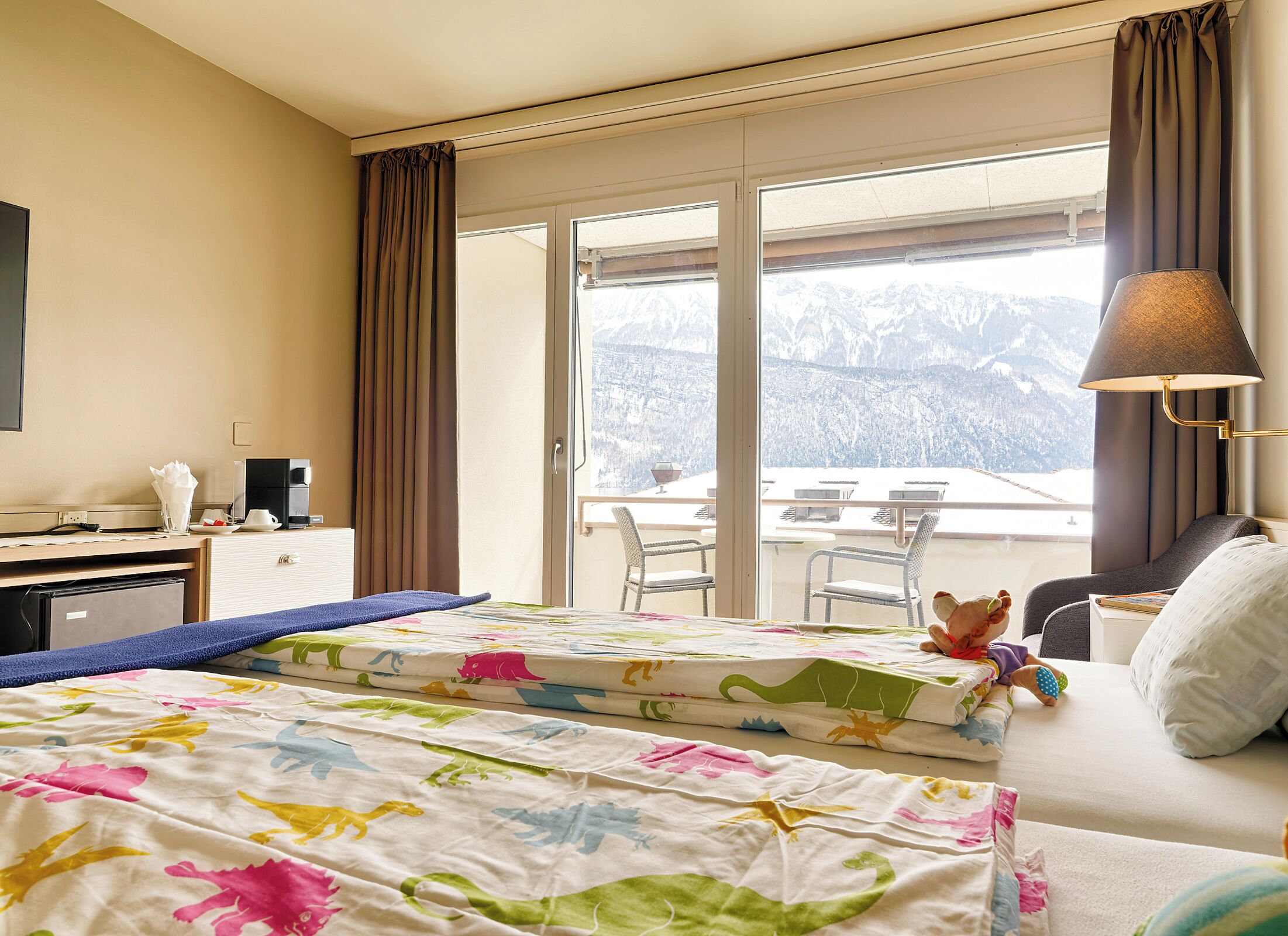 Een hotelkamer met een bed, een bureau, een stoel, een lamp en uitzicht op bergen door het raam.