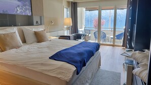Hotellihuone, jossa on suuri sänky, työpöytä kannettavalla tietokoneella, tuoli ja parveke, jolta on näkymä vuorille.
