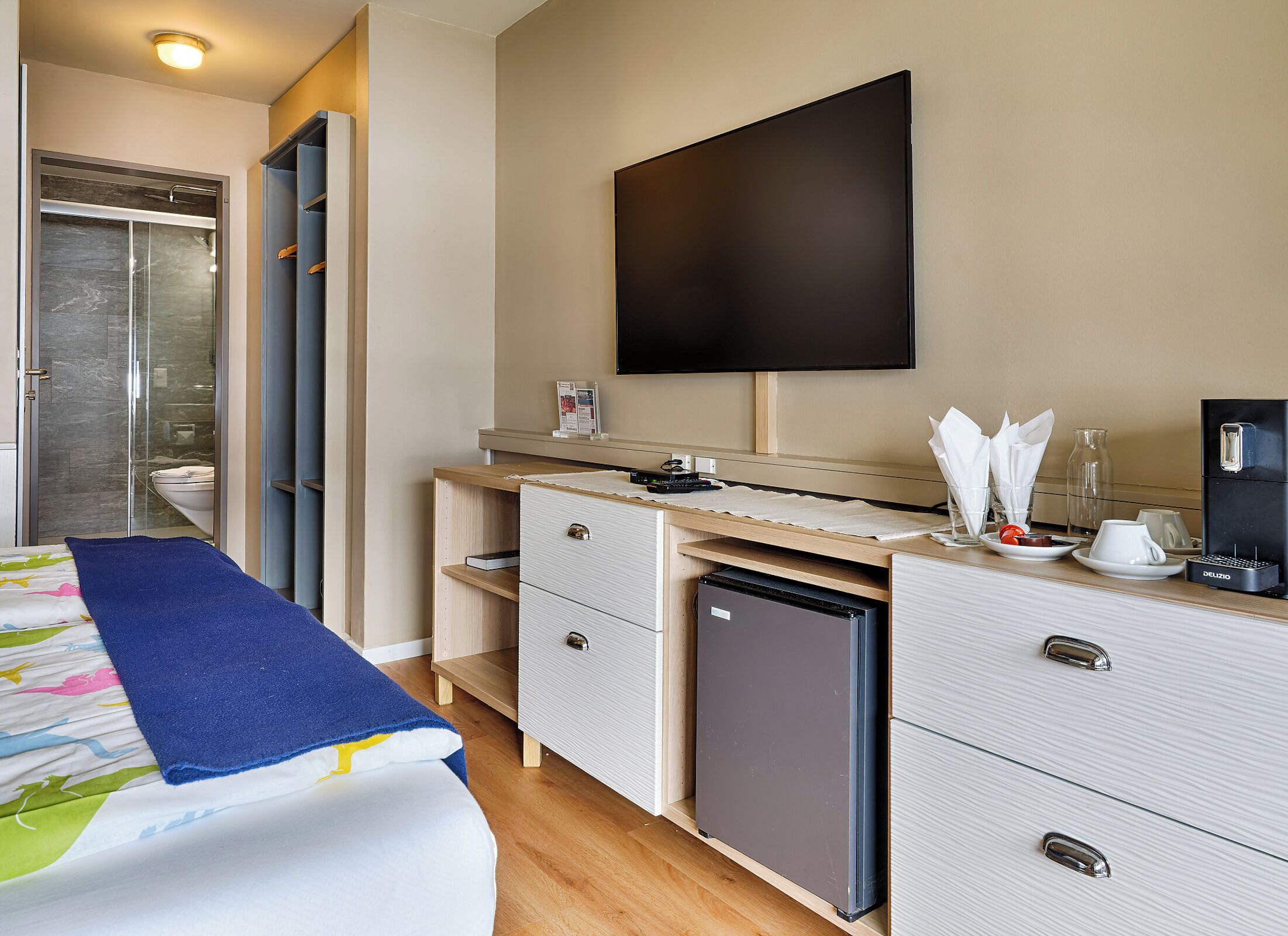 A modern hotel room with a shower, a bed with a blue blanket, a desk with a coffee machine, and a flat-screen TV.